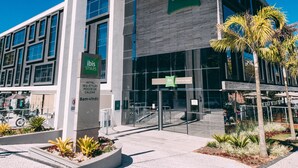 Un hotel moderno con un llamativo letrero de 'ibis Styles', un acceso cubierto y una plaza pavimentada con jardines.
