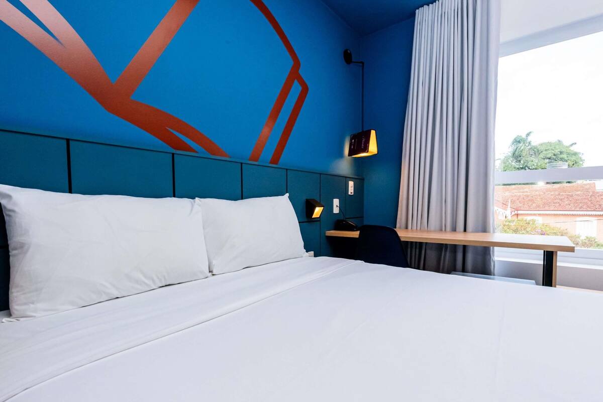 Habitación de hotel con una cama grande, un escritorio, una silla, una lámpara y una ventana con cortinas.