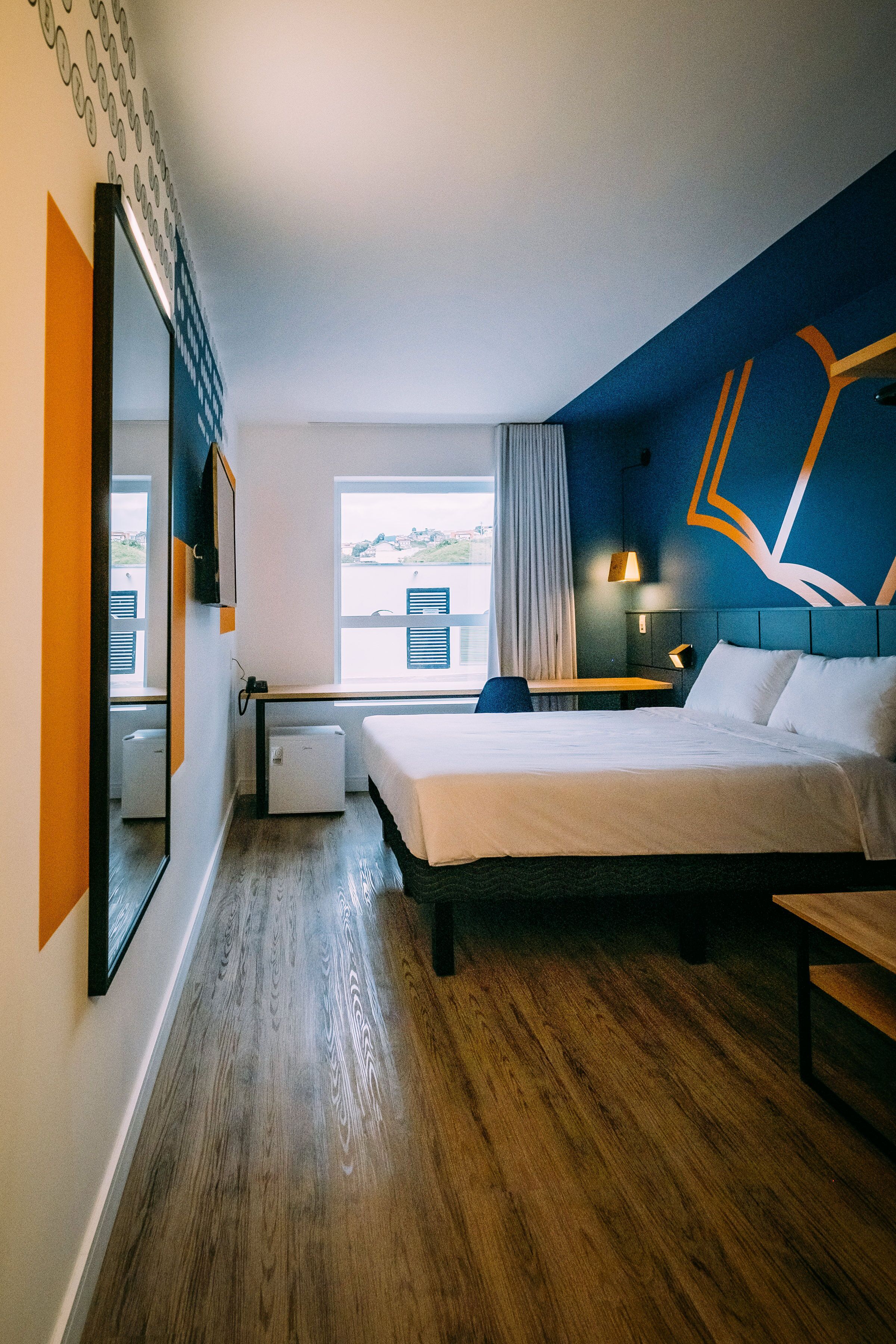 A modern hotel room with a large bed, a desk, a chair, and a window with a view of the outdoors.