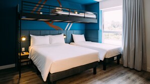 A room with two beds, a bunk bed, wooden flooring, a window with curtains, and a nightstand with a lamp.