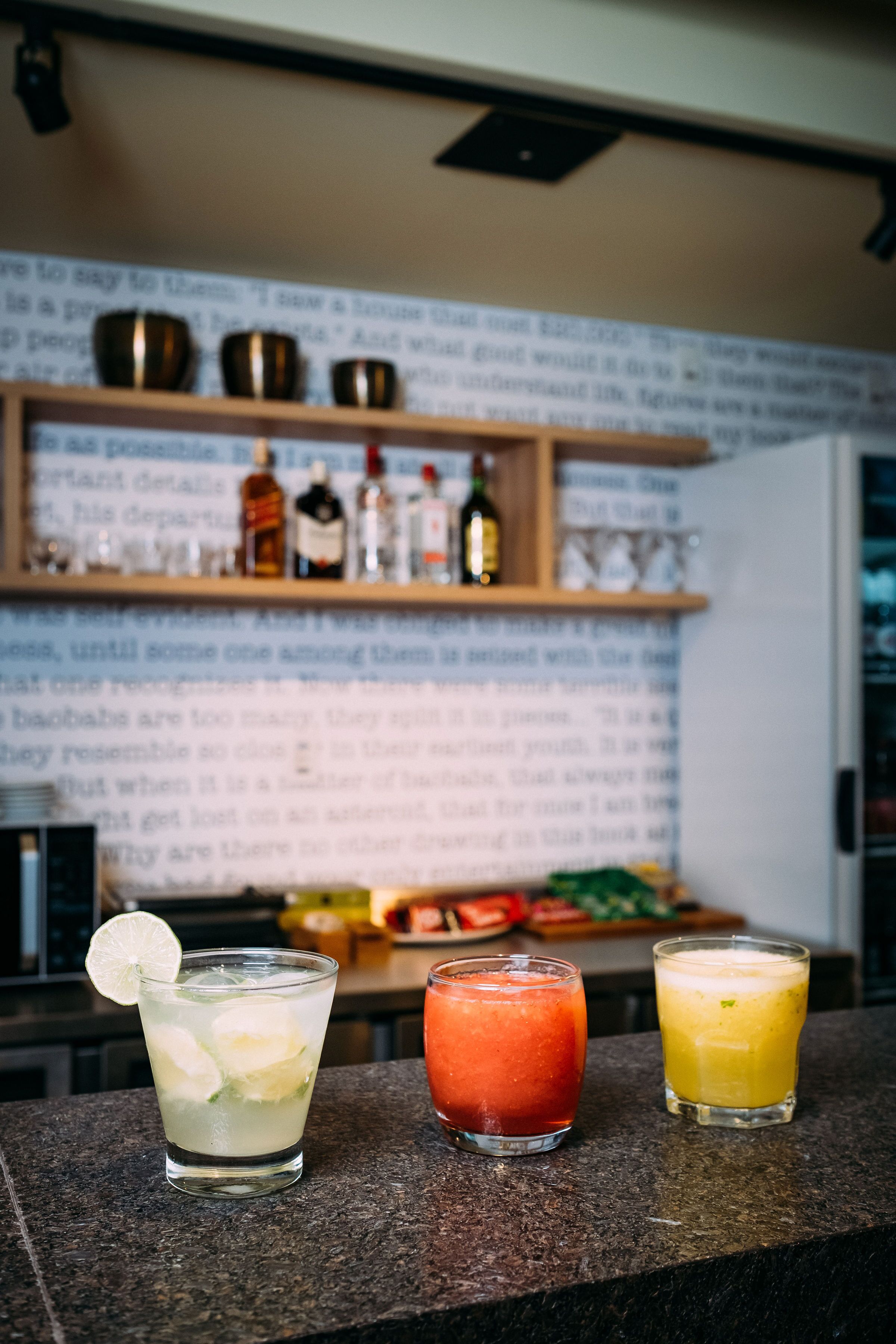 Three glasses of mixed drinks on a counter with a blurred background of shelves and bottles.