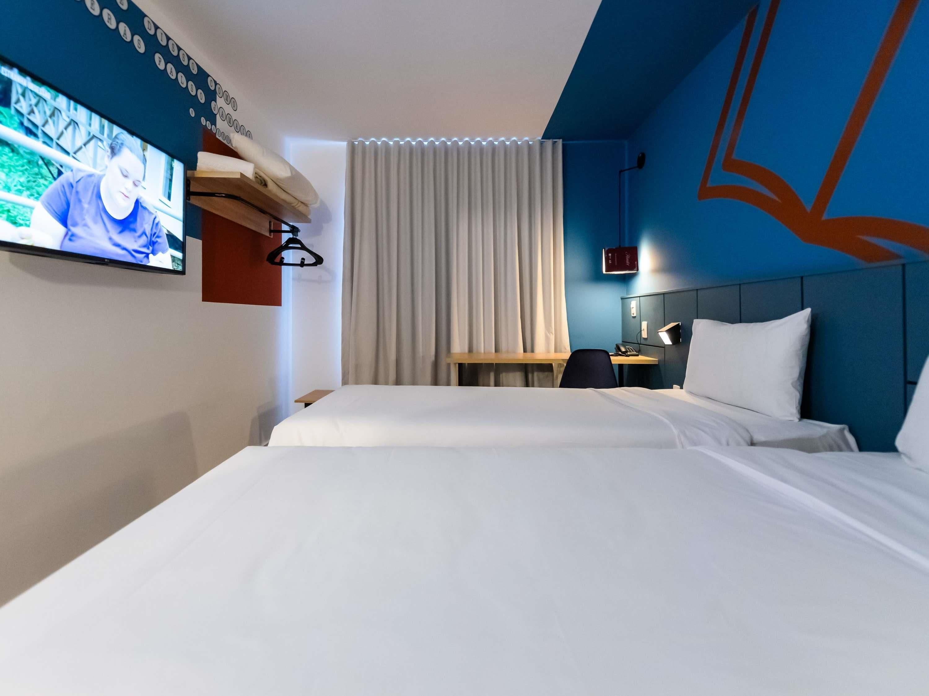 A hotel room with a bed, a desk, a TV mounted on the wall, and a window with curtains.