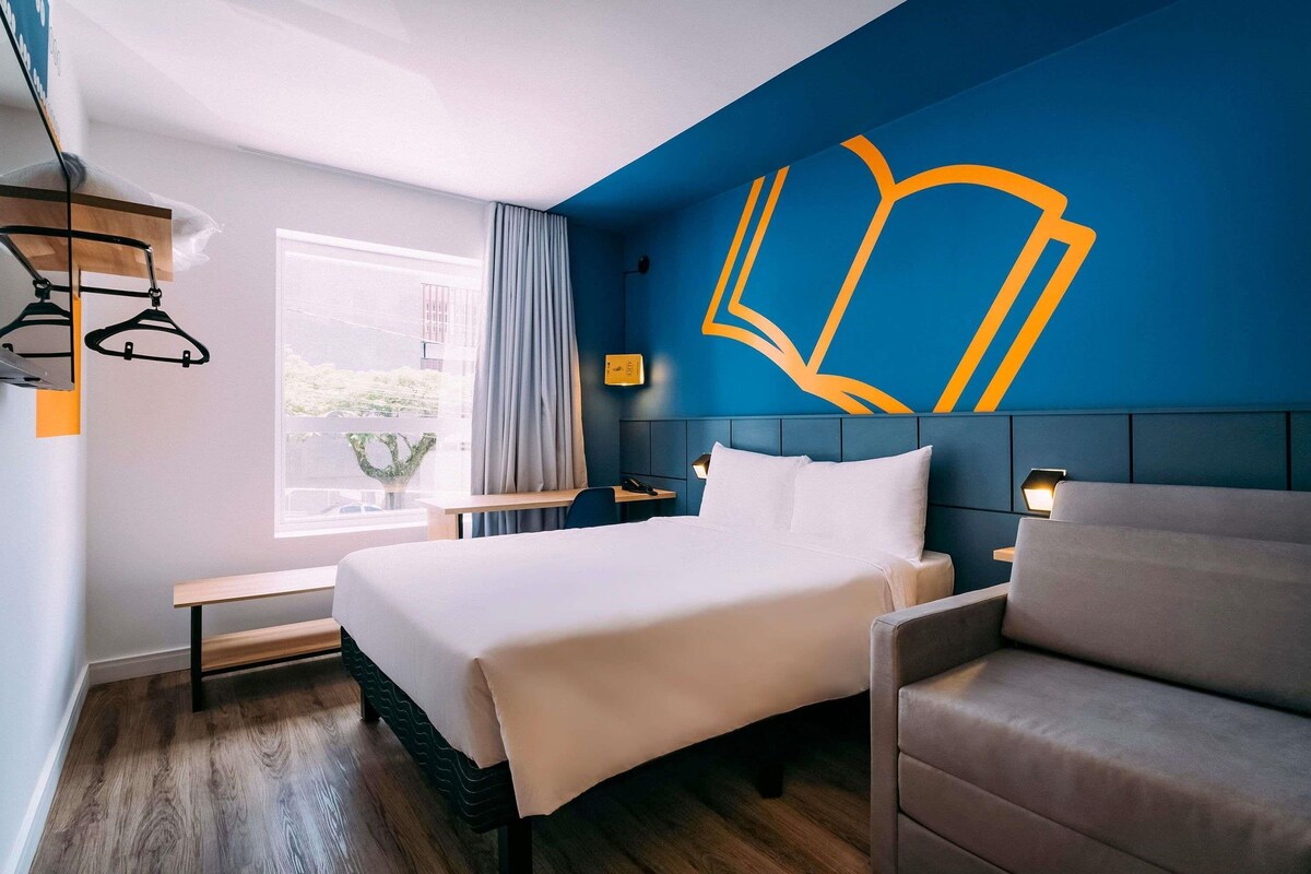 Habitación de hotel con una cama grande, un escritorio, una silla y una ventana con cortinas.