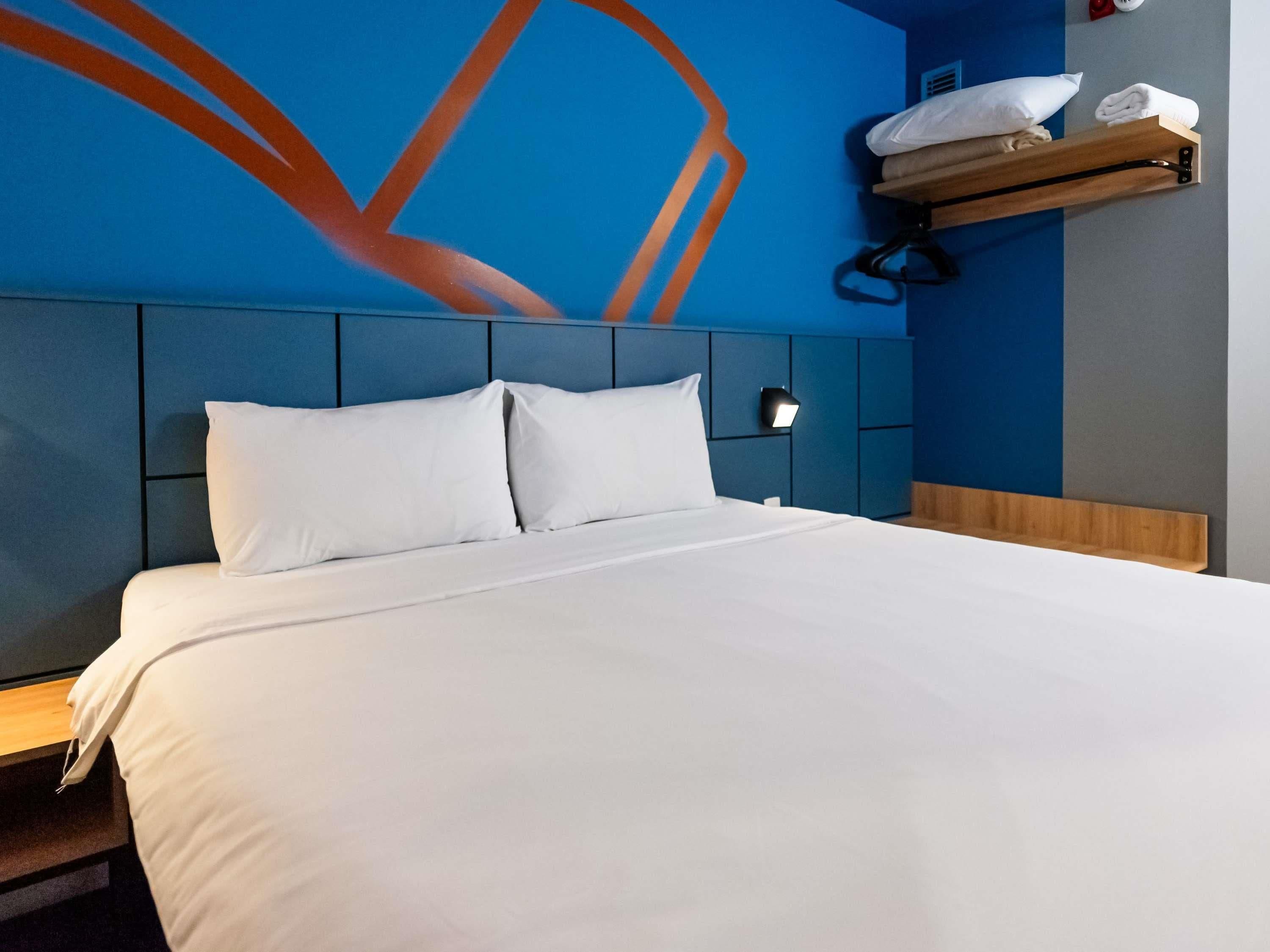 A hotel room with a large bed, a headboard with a geometric design, a wall-mounted shelf with towels, and a wall art piece.