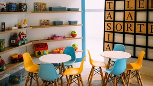 Zona de juegos infantiles con juguetes de colores, una mesa blanca con sillas azules y amarillas, y una pared con letras grandes que deletrean una palabra.