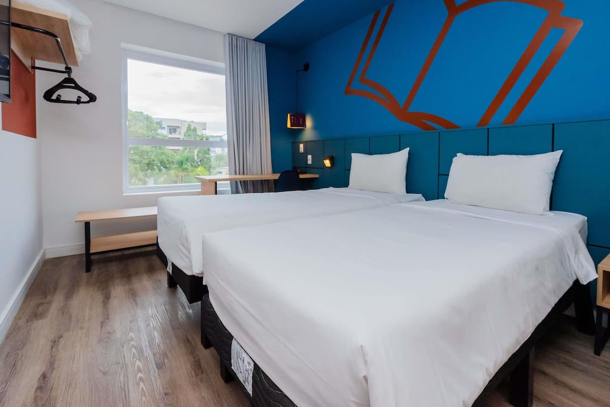 A hotel room with two beds, a large window, a view of greenery, a blue accent wall, and a hanging coat rack.