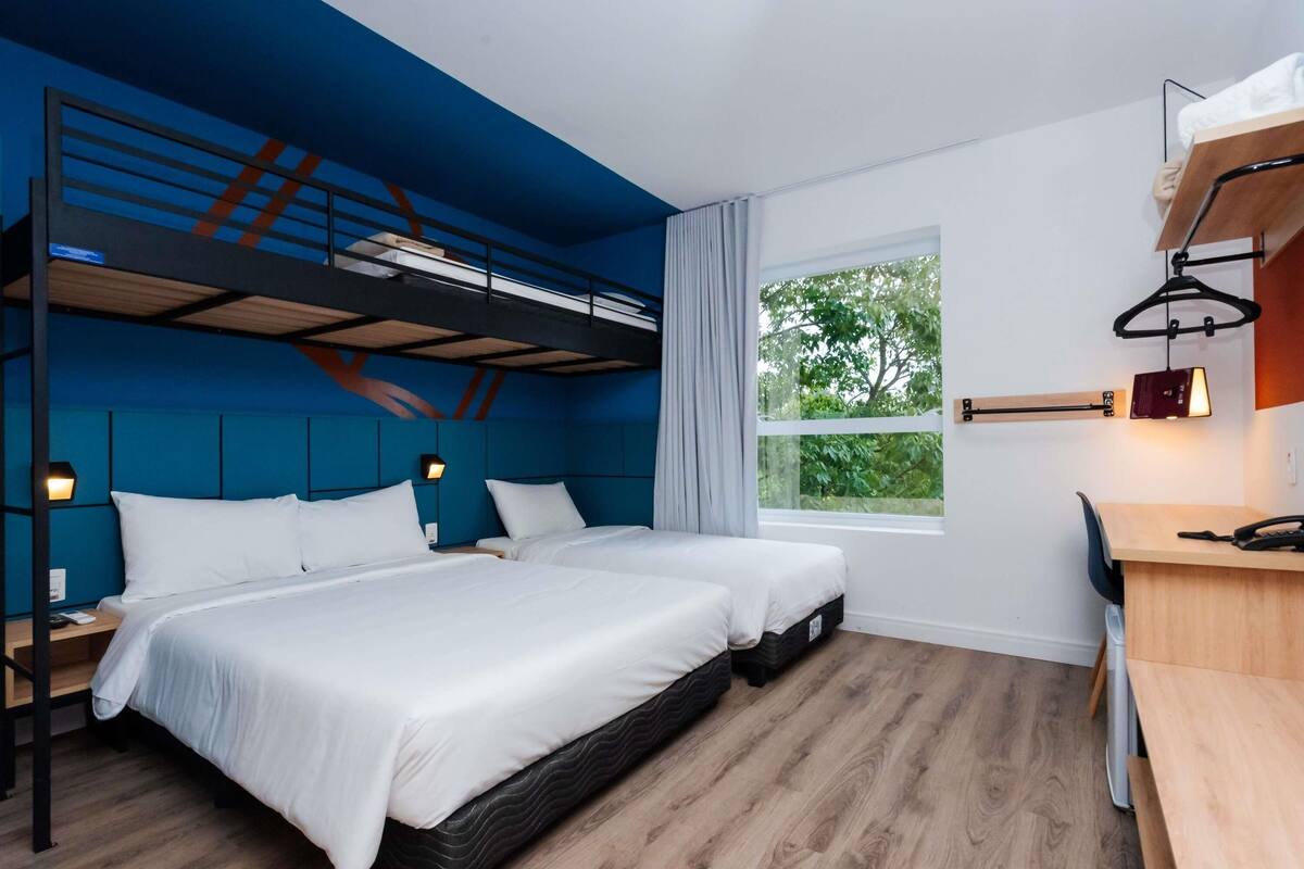 Habitación de hotel moderna con dos camas, un escritorio, una silla, un ventanal con vistas a zonas verdes y una litera encima de las camas.