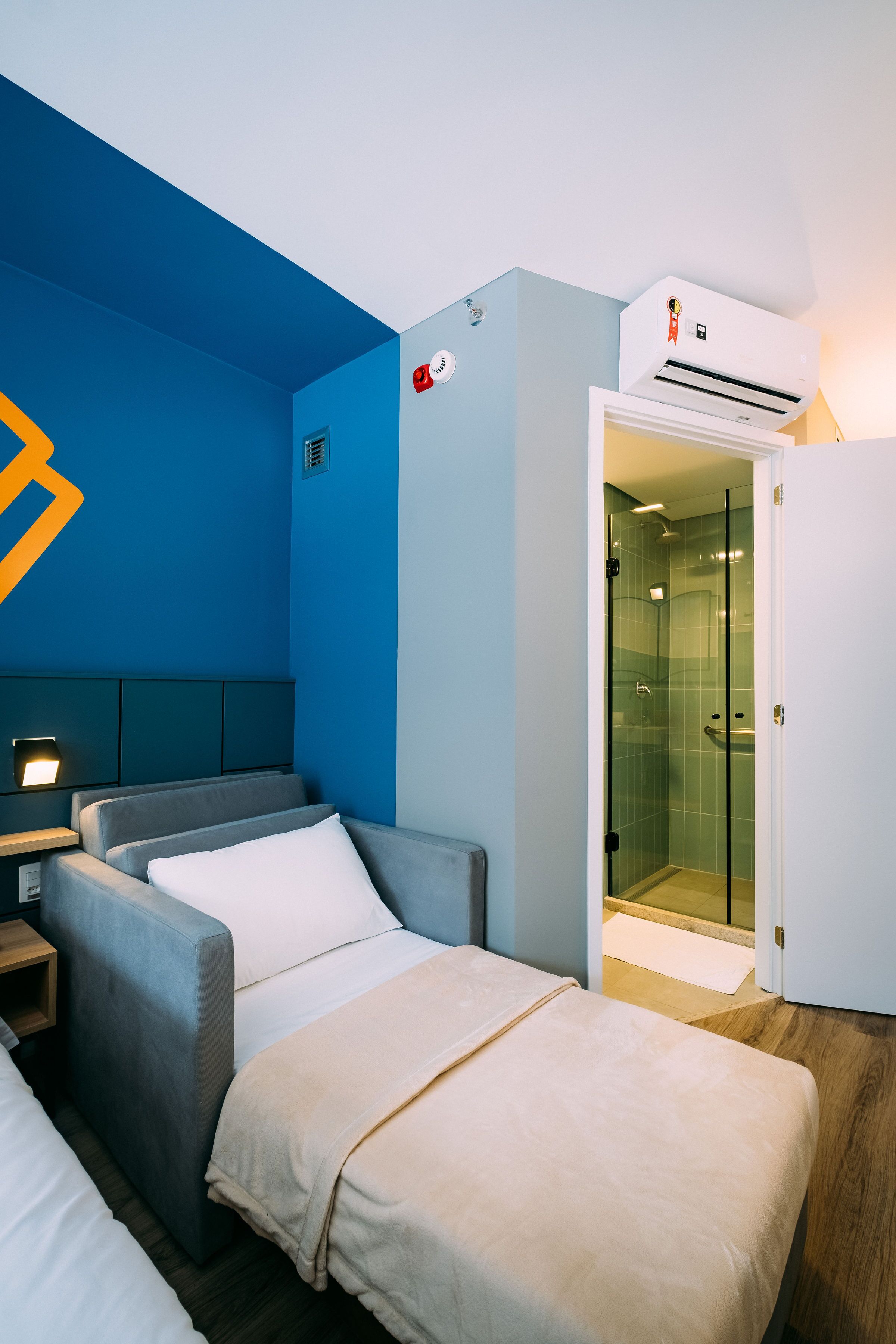 A hotel room with a blue accent wall, a bed with white bedding, a grey sofa, a bedside table with a lamp, and a glass-enclosed shower area.