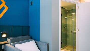 A hotel room with a blue accent wall, a bed with white bedding, a grey sofa, a bedside table with a lamp, and a glass-enclosed shower area.