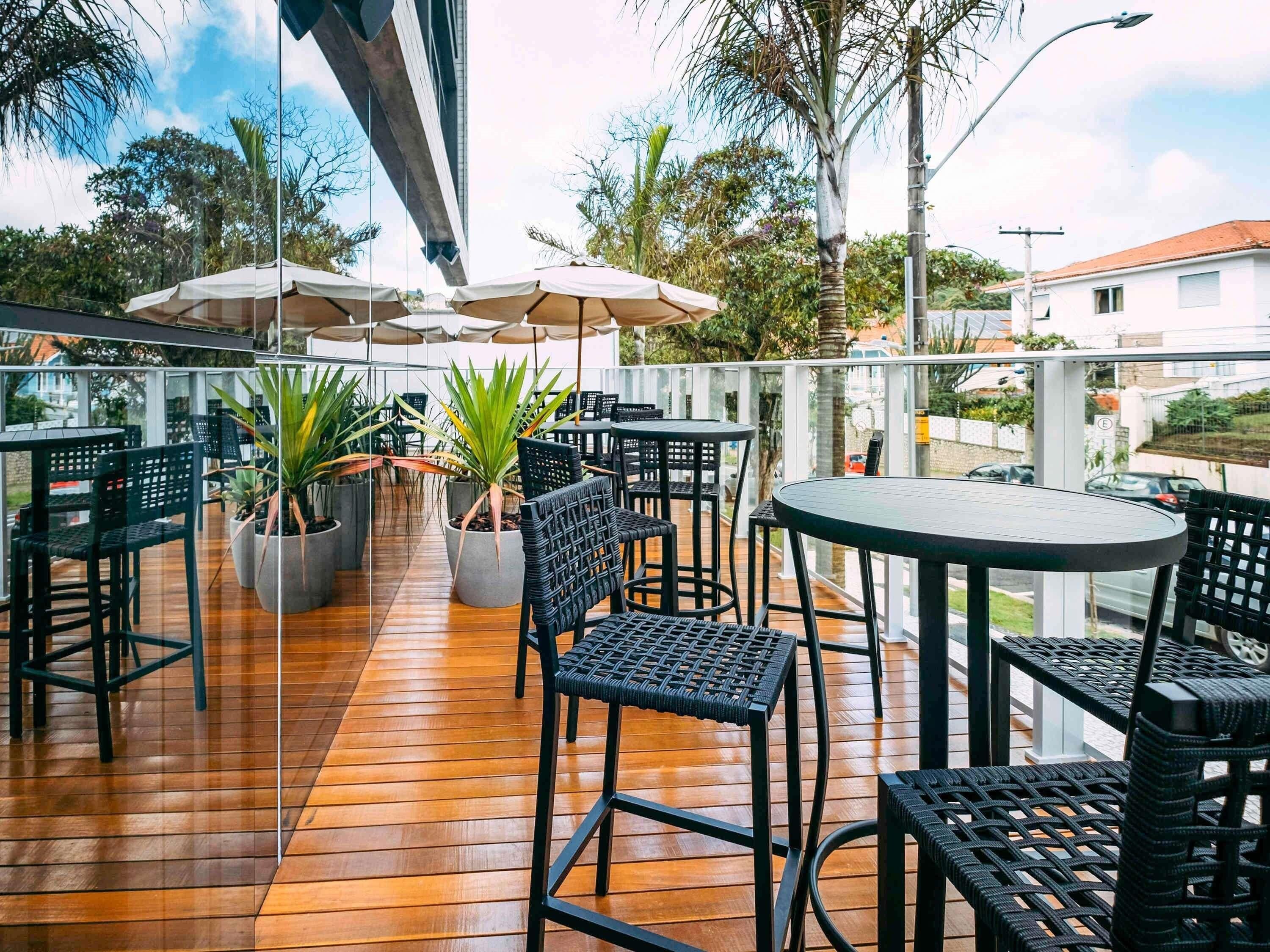 An outdoor patio with wooden flooring, black wicker chairs, and tables. There are umbrellas providing shade, and potted plants are placed around the seating area.