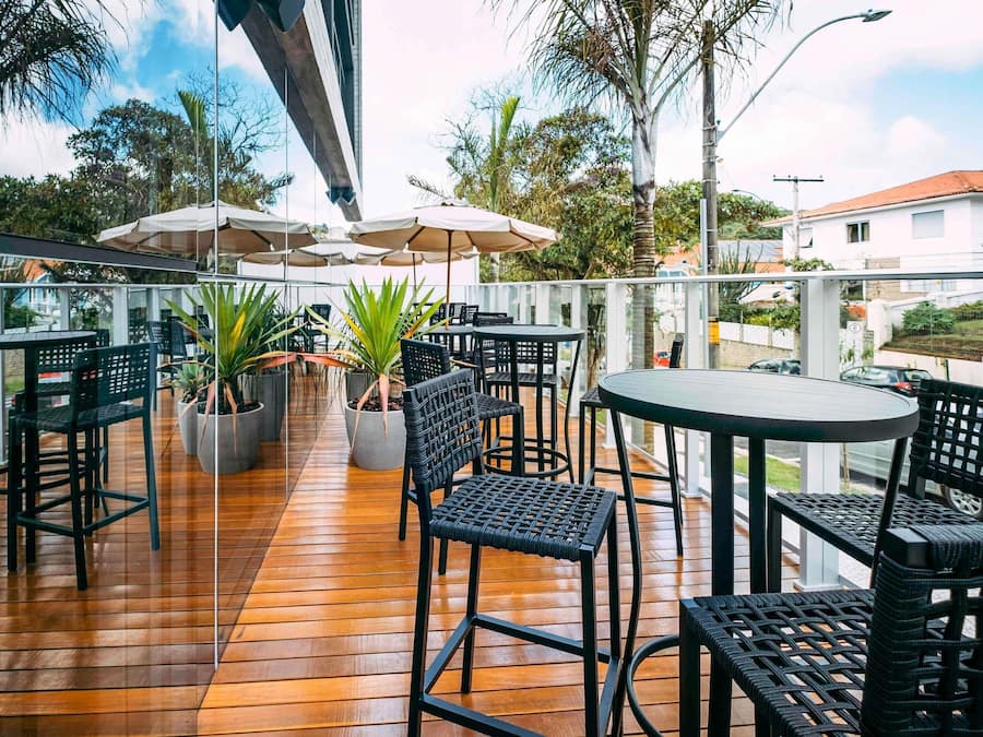 An outdoor patio with wooden flooring, black wicker chairs, and tables. There are umbrellas providing shade, and potted plants are placed around the seating area.