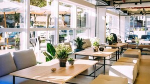 Un espacio interior moderno con mesas, sillas, ventanas amplias y plantas en macetas.