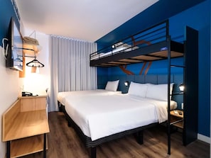 Quarto de hotel com uma cama beliche, uma escrivaninha com cadeira, uma mesinha de cabeceira e uma televisão fixada na parede.