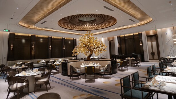 A modern hotel lobby with a central seating area, multiple dining tables, and a distinctive ceiling design.