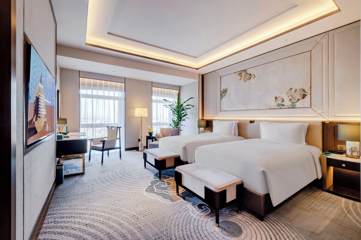A hotel room with two beds, a desk, a chair, a small table, a lamp, and a large painting on the wall.