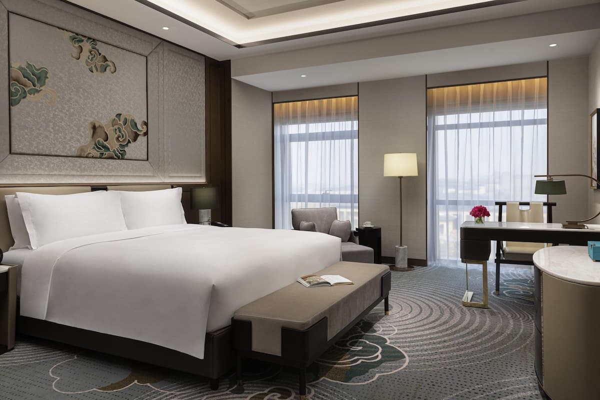 A hotel room with a large bed, a desk, a chair, and a bench. There are two windows with curtains, a lamp, and a painting on the wall.