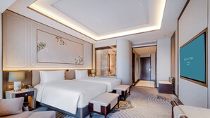 A modern hotel room with a large bed, bedside tables, a seating area, and a large screen displaying the hotel name.