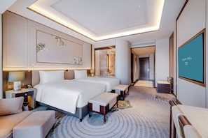 A modern hotel room with a large bed, bedside tables, a seating area, and a large screen displaying the hotel name.