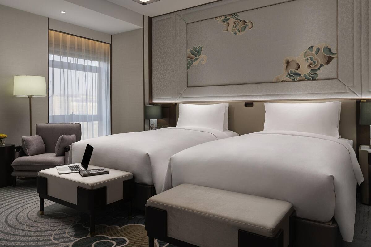 A hotel room with two beds, a desk, a chair, a lamp, and a large decorative wall panel.