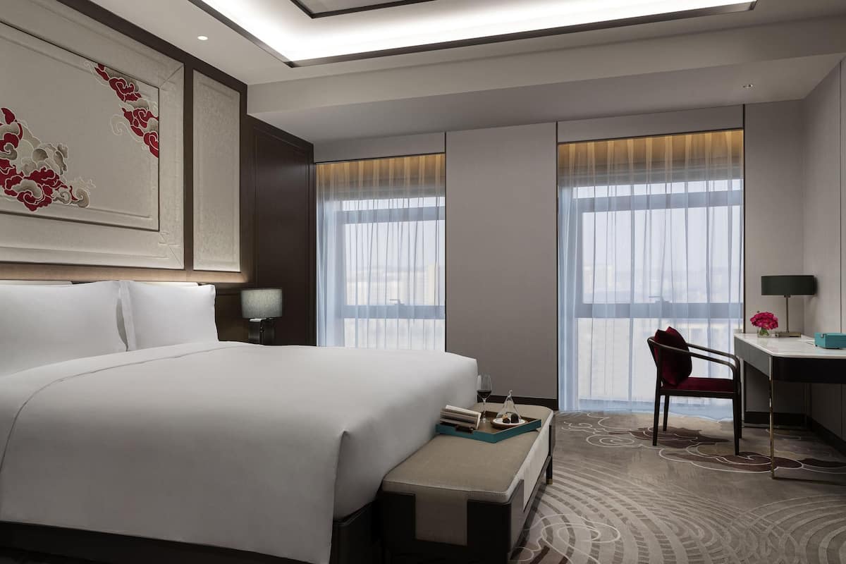 A hotel room with a large bed, a desk, and two windows with sheer curtains.