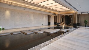 A modern indoor swimming pool with a minimalist design, featuring a marble wall, a wooden reception desk, and a large circular mirror.