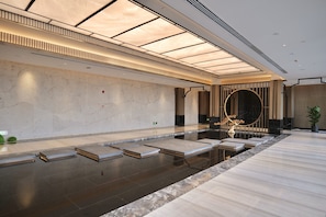 A modern indoor swimming pool with a minimalist design, featuring a marble wall, a wooden reception desk, and a large circular mirror.