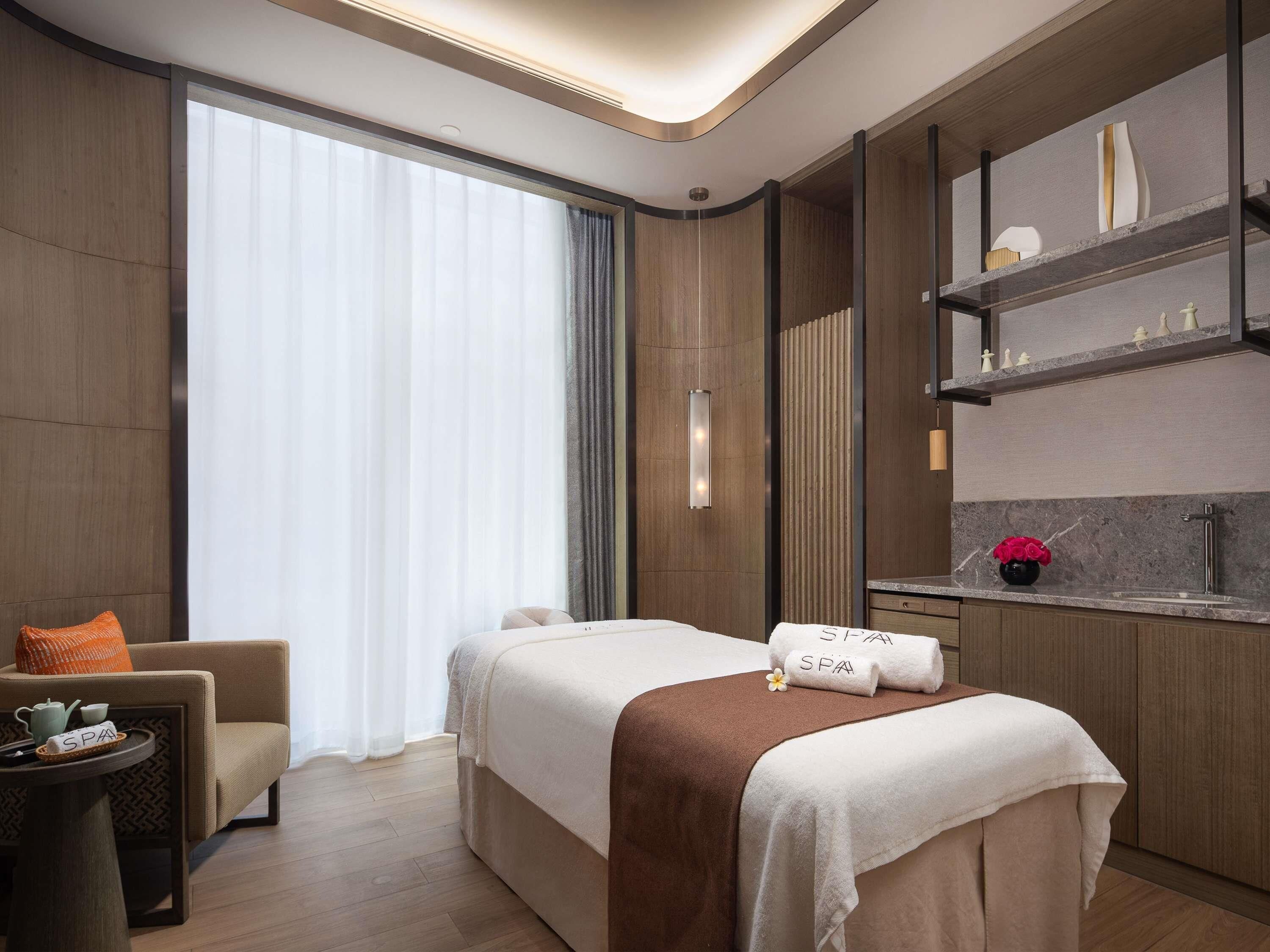 A modern hotel room with a bed, a seating area, a vanity with a mirror, and a large window with curtains.