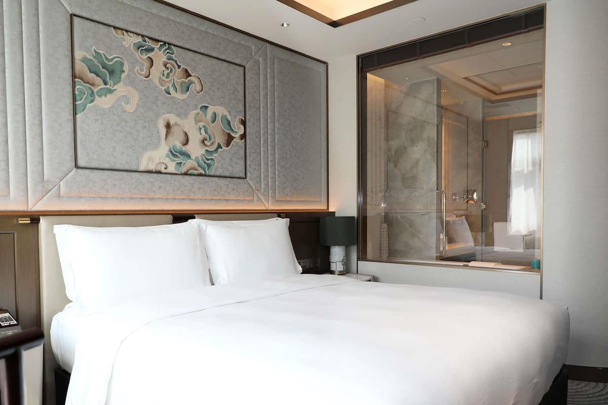 A hotel room with a large bed, a wall-mounted artwork, a glass shower enclosure, and a nightstand with a lamp.