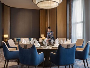 A hotel dining area with a round table set for four, blue upholstered chairs, and a staff member attending to the table.