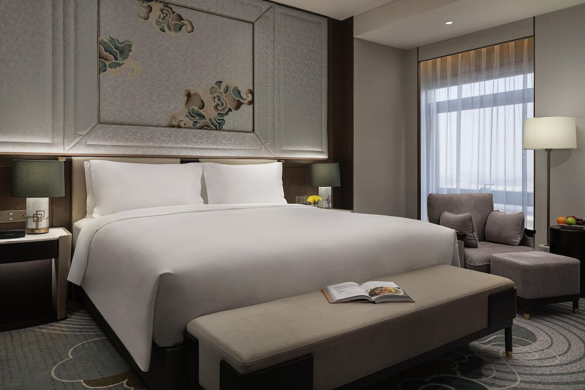 A hotel room with a large bed, a bench, a chair, a bedside table, a lamp, and a window with curtains.