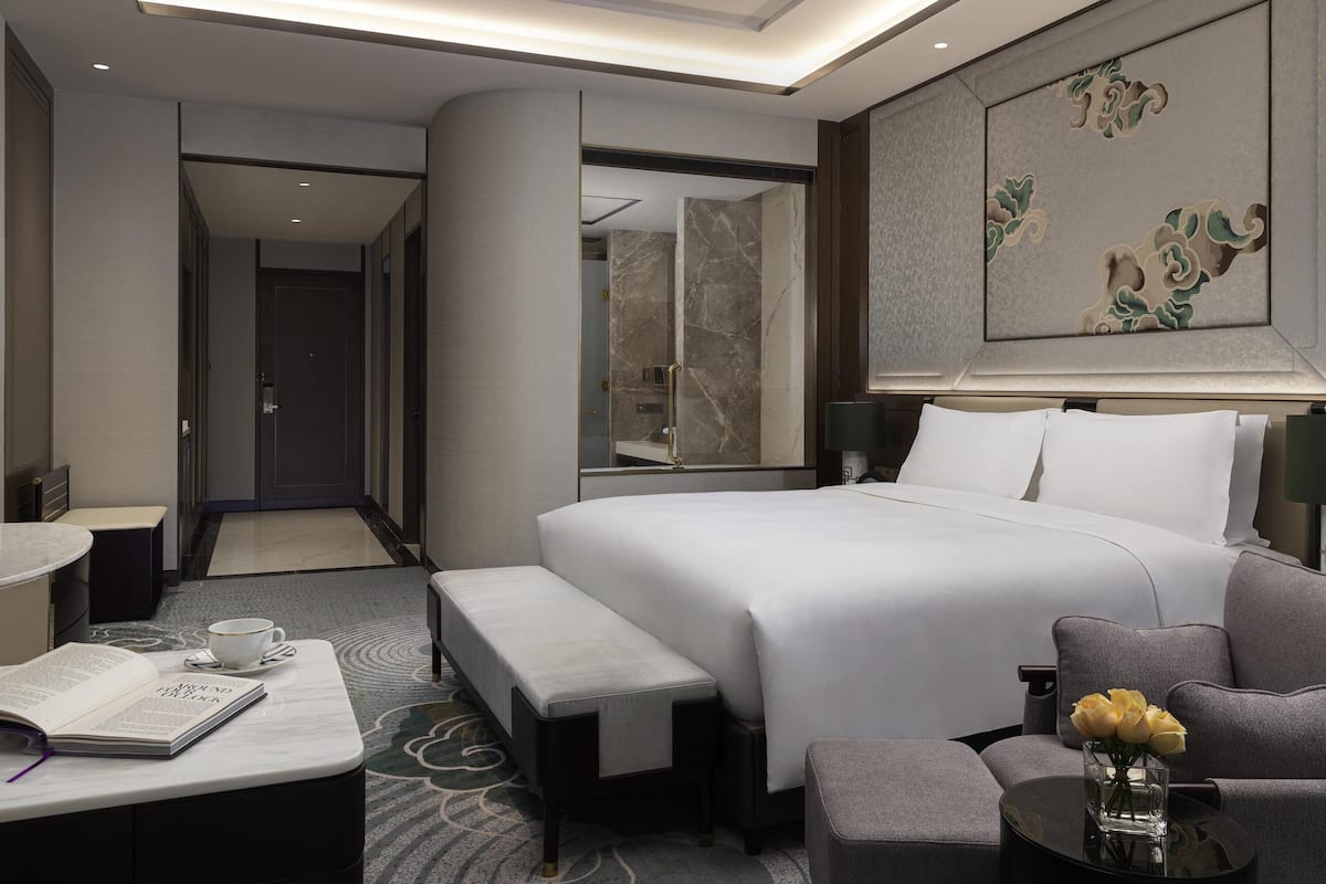 A modern hotel room with a large bed, a seating area with a bench, a small table with a cup and book, and a decorative wall piece.