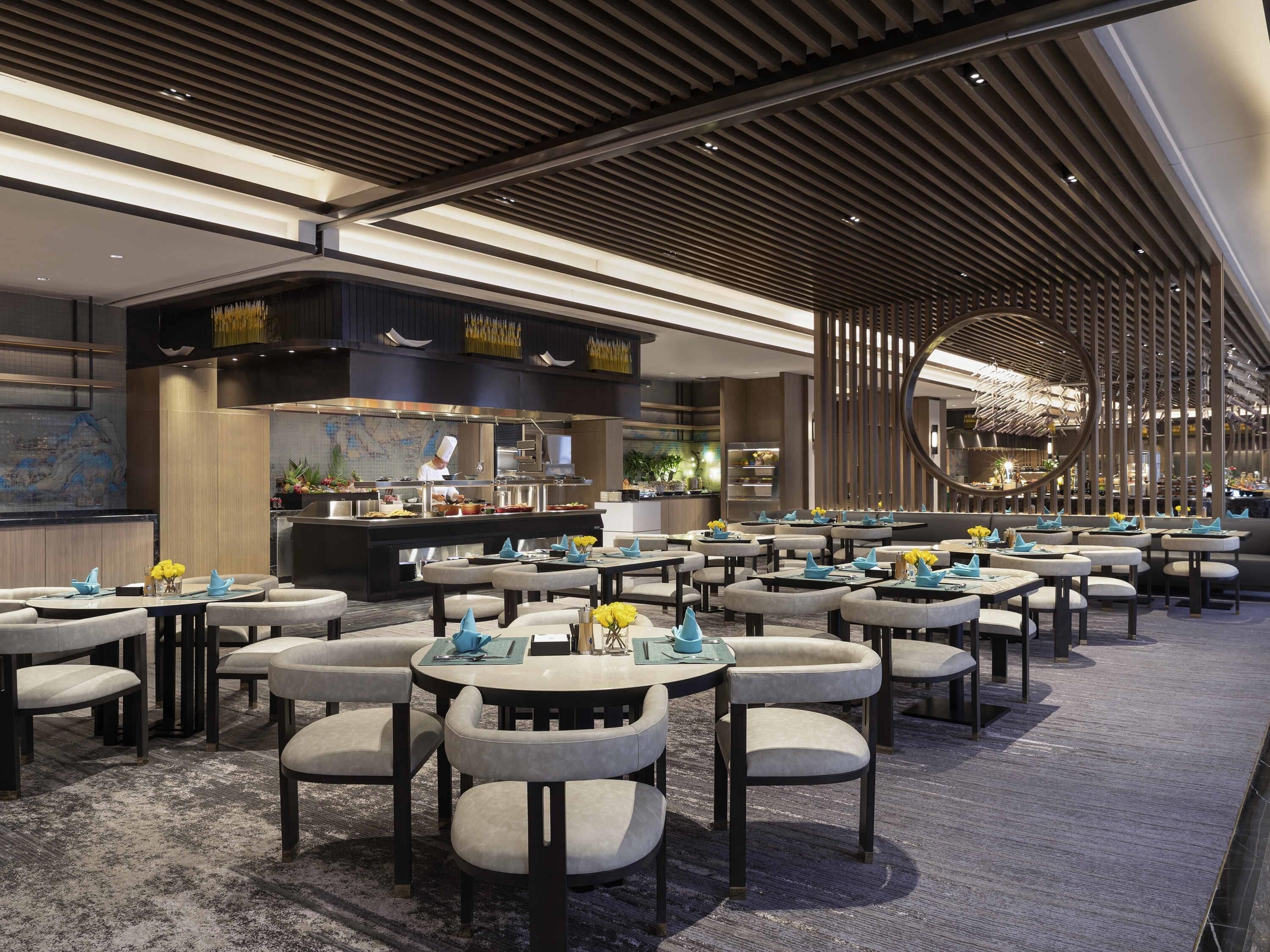 A modern restaurant with round tables and chairs, a chef preparing food, and a decorative ceiling.