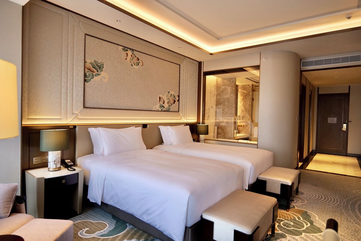 A hotel room with two beds, a nightstand with a lamp, a mirror, and a carpeted floor.