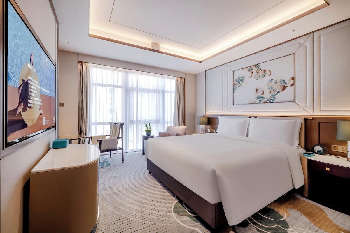 A hotel room with a large bed, a desk, a chair, a TV, and a window with curtains.
