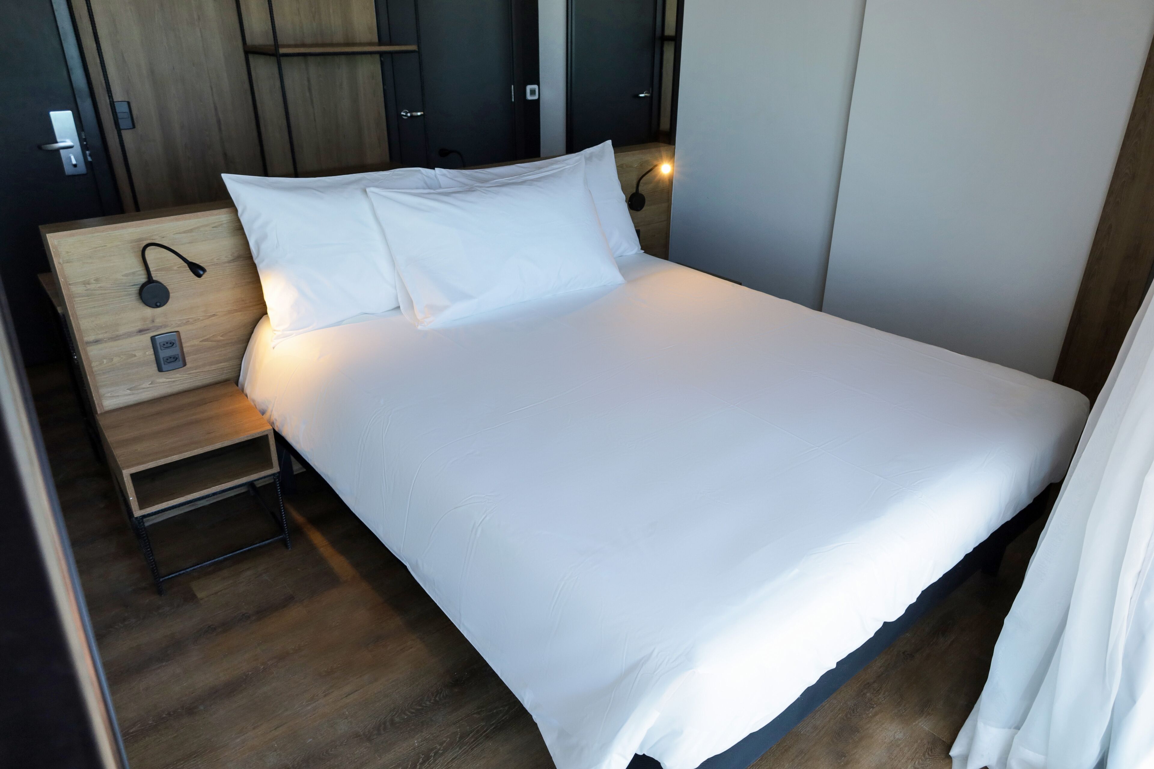 A neatly made bed with white linens, a bedside table with a lamp, and a wooden nightstand.