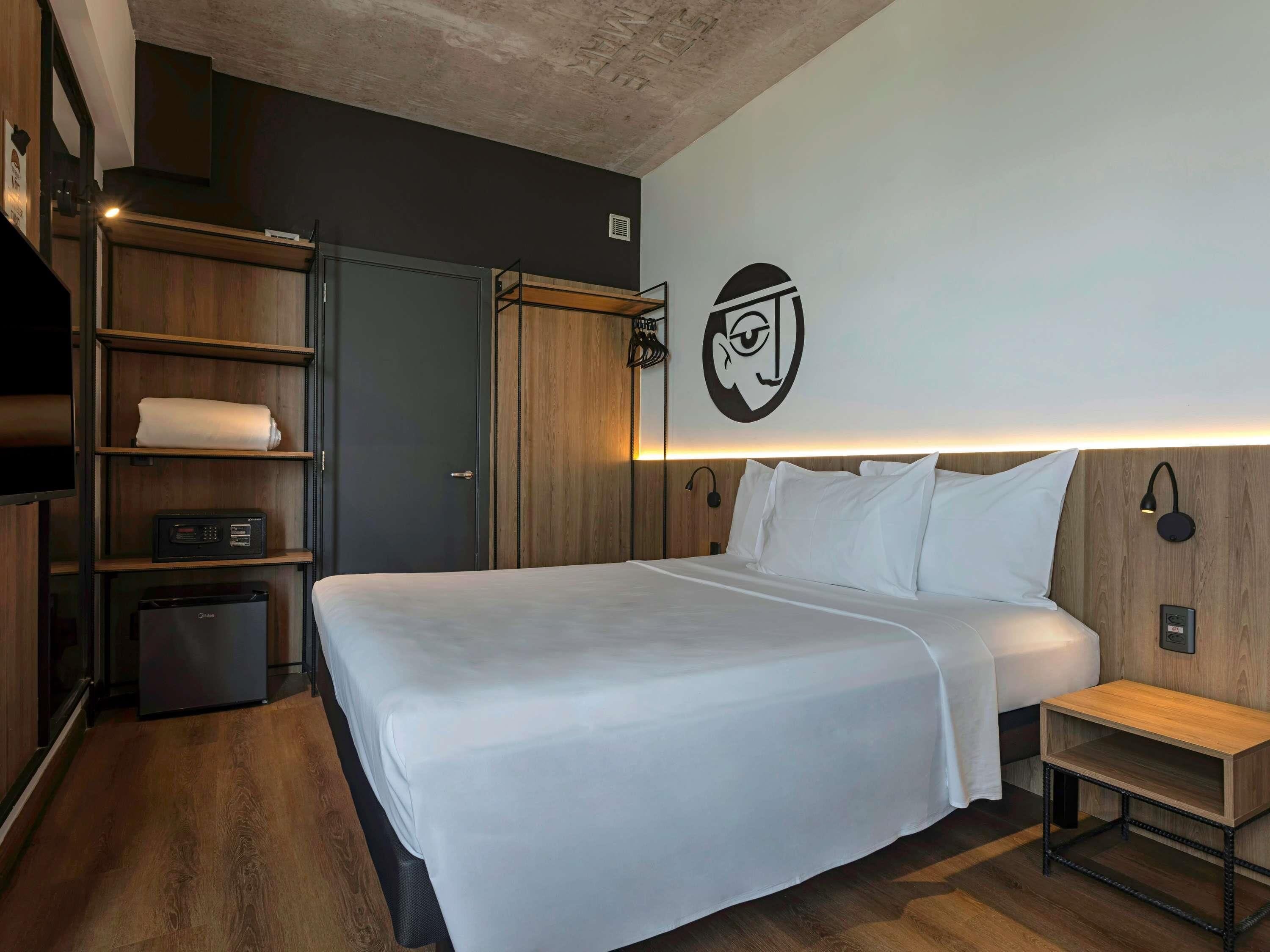 Una habitación de hotel moderna con una cama grande, un armario de madera, estanterías empotradas y una lámpara fijada a la pared.