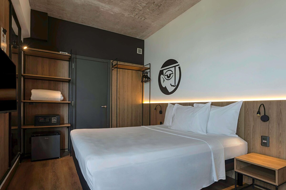 Una habitación de hotel moderna con una cama grande, un armario de madera, estanterías empotradas y una lámpara fijada a la pared.