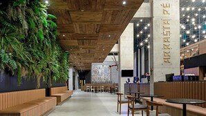 Un interior moderno con paneles de techo de madera, plantas en una pared verde y una zona de estar con mesas y sillas.