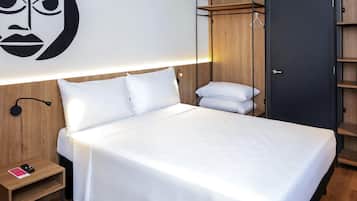 Una cama con ropa blanca, cabecera de madera, mesita de noche con teléfono, lámpara de pared y armario con perchero.
