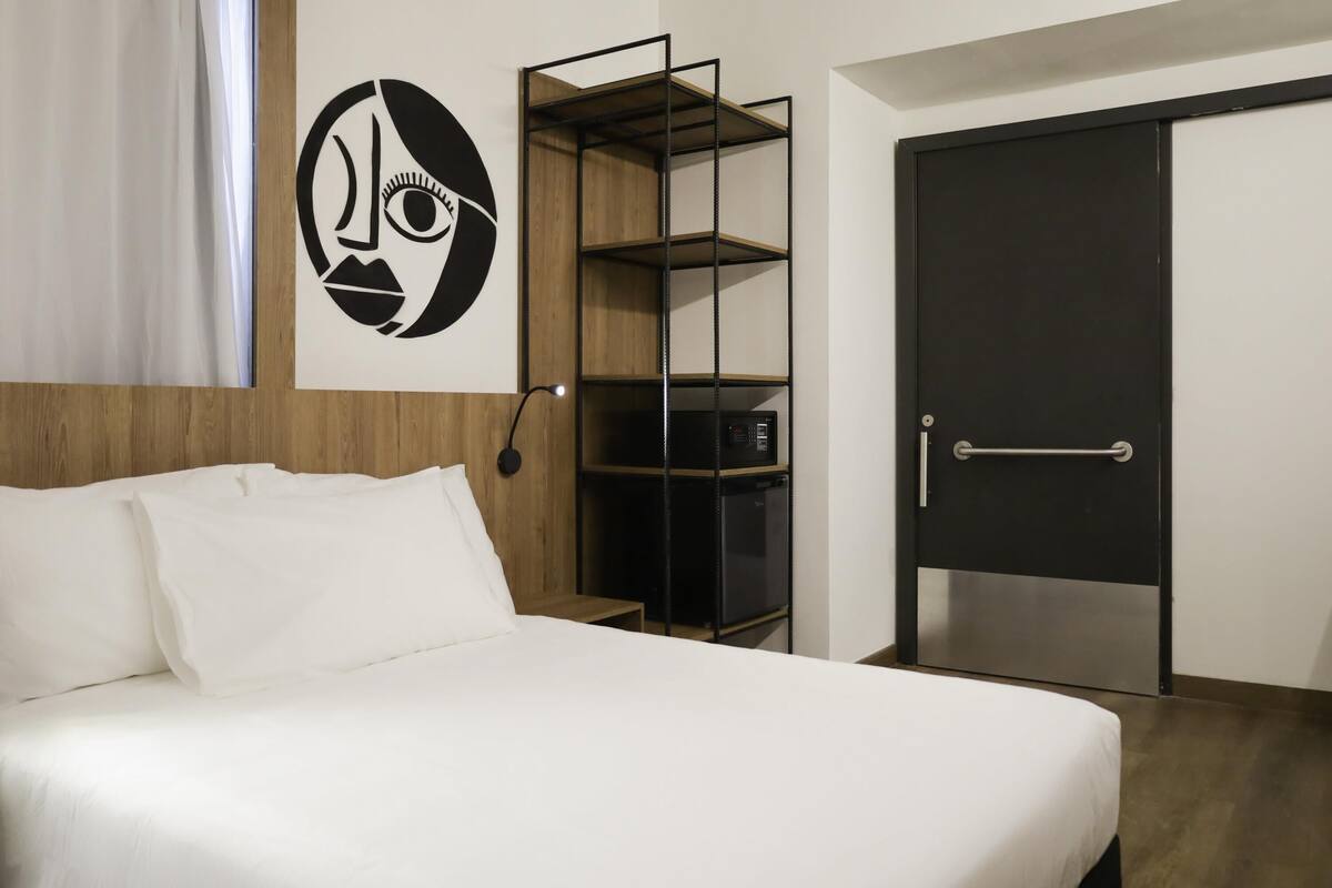 A hotel room with a bed, a wall-mounted artwork, a built-in shelf unit, and a door.