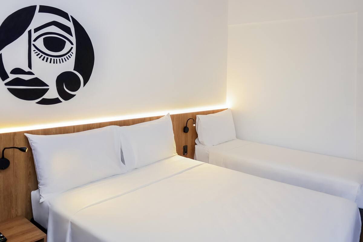 A hotel room with two single beds, a wooden headboard, a wall-mounted light, and a nightstand with a remote control.