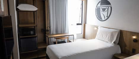 A hotel room with a bed, a desk, a wardrobe, and a wall-mounted light fixture.