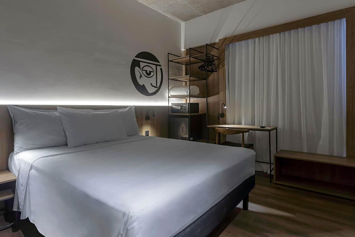 A modern hotel room with a large bed, a desk, a chair, a wardrobe, and a wall-mounted shelf.