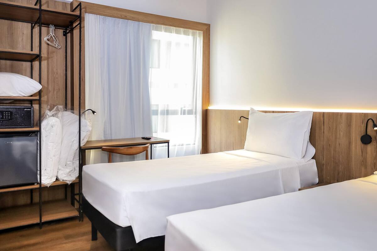 A hotel room with two beds, a desk, a chair, a wardrobe, and a window with curtains.