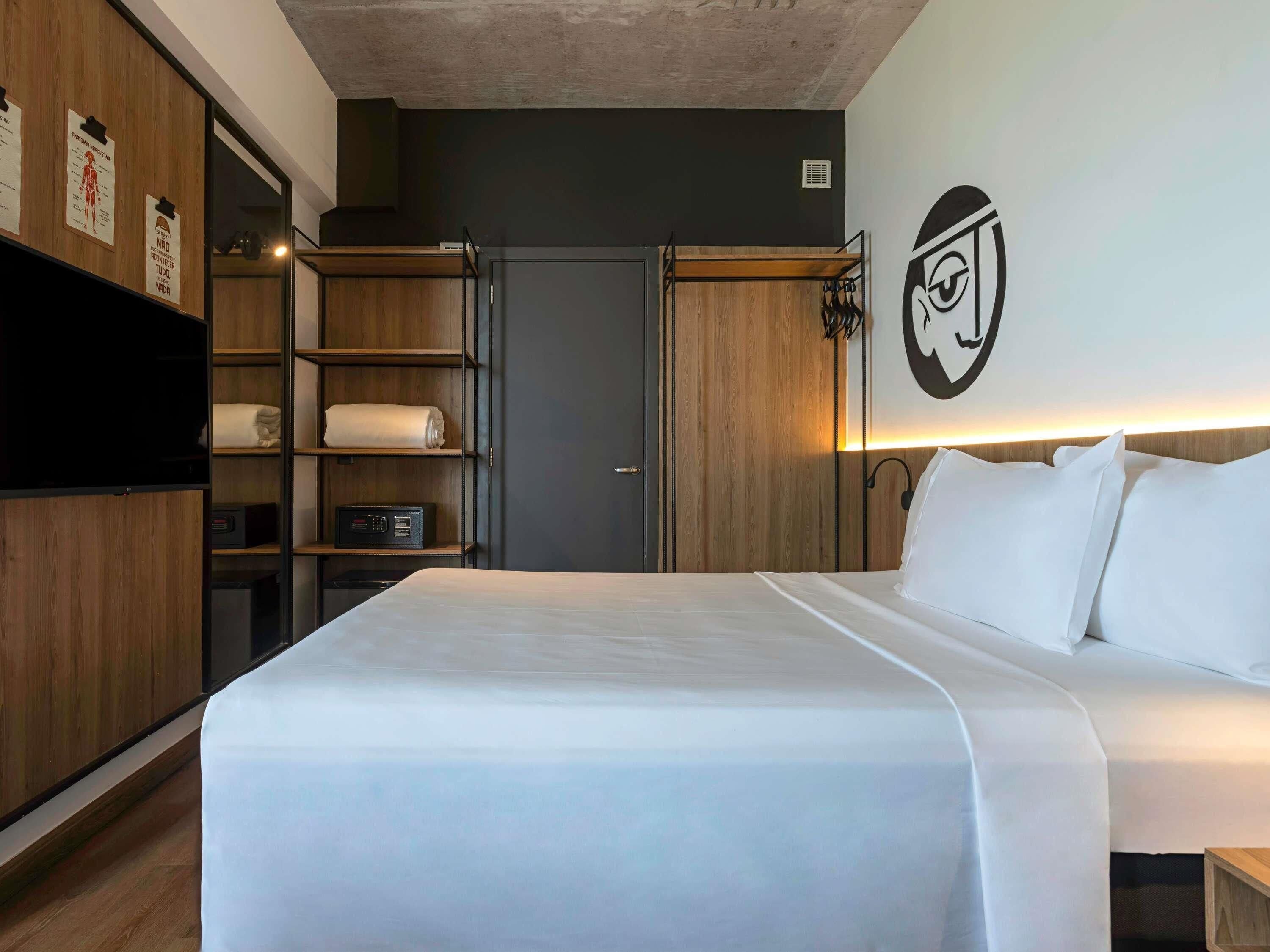 A hotel room with a bed, wooden wardrobe, shelves, a flat-screen TV, and a wall-mounted artwork.