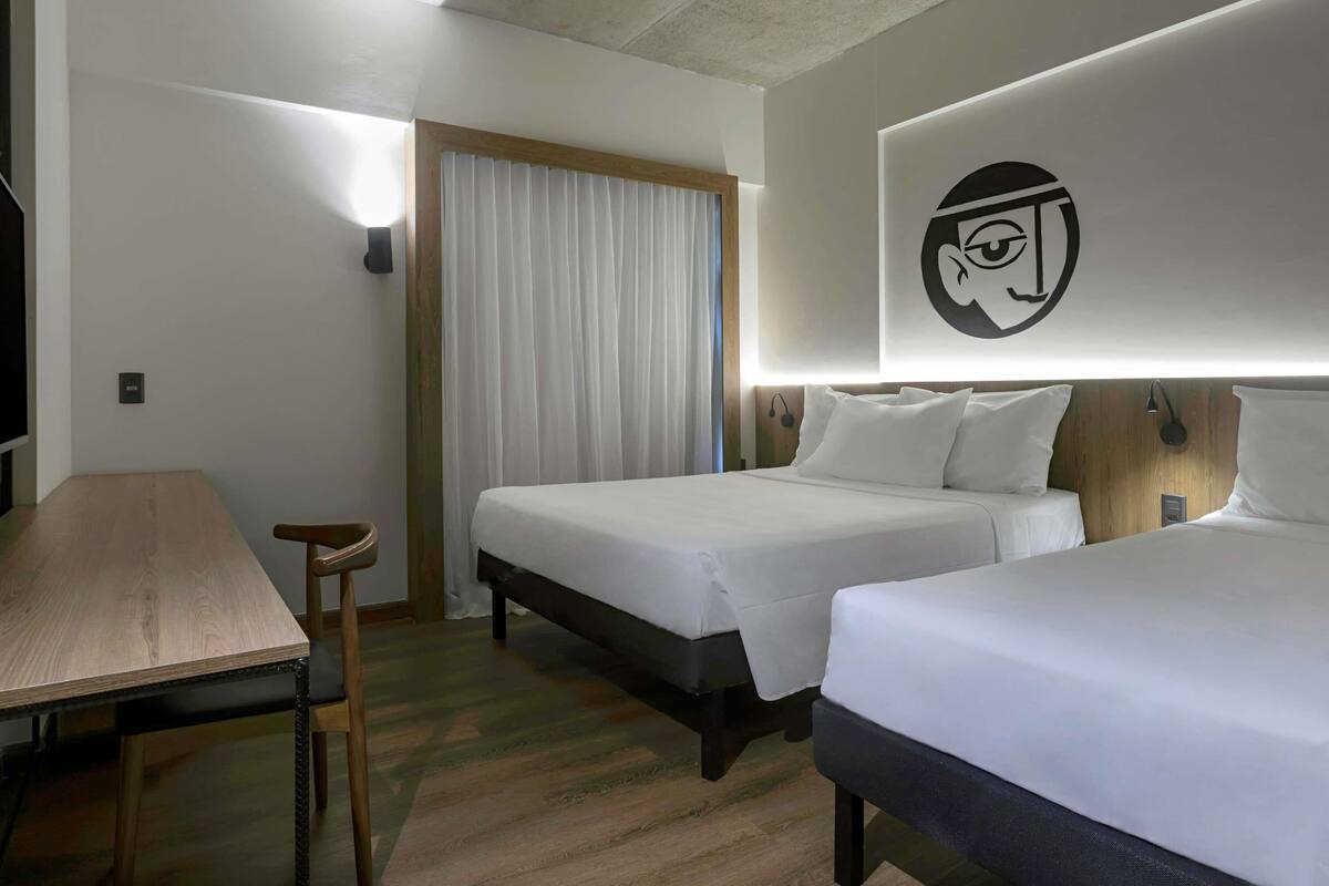 Habitación de hotel con dos camas, un escritorio, una silla, una cortina y una lámpara fijada a la pared.