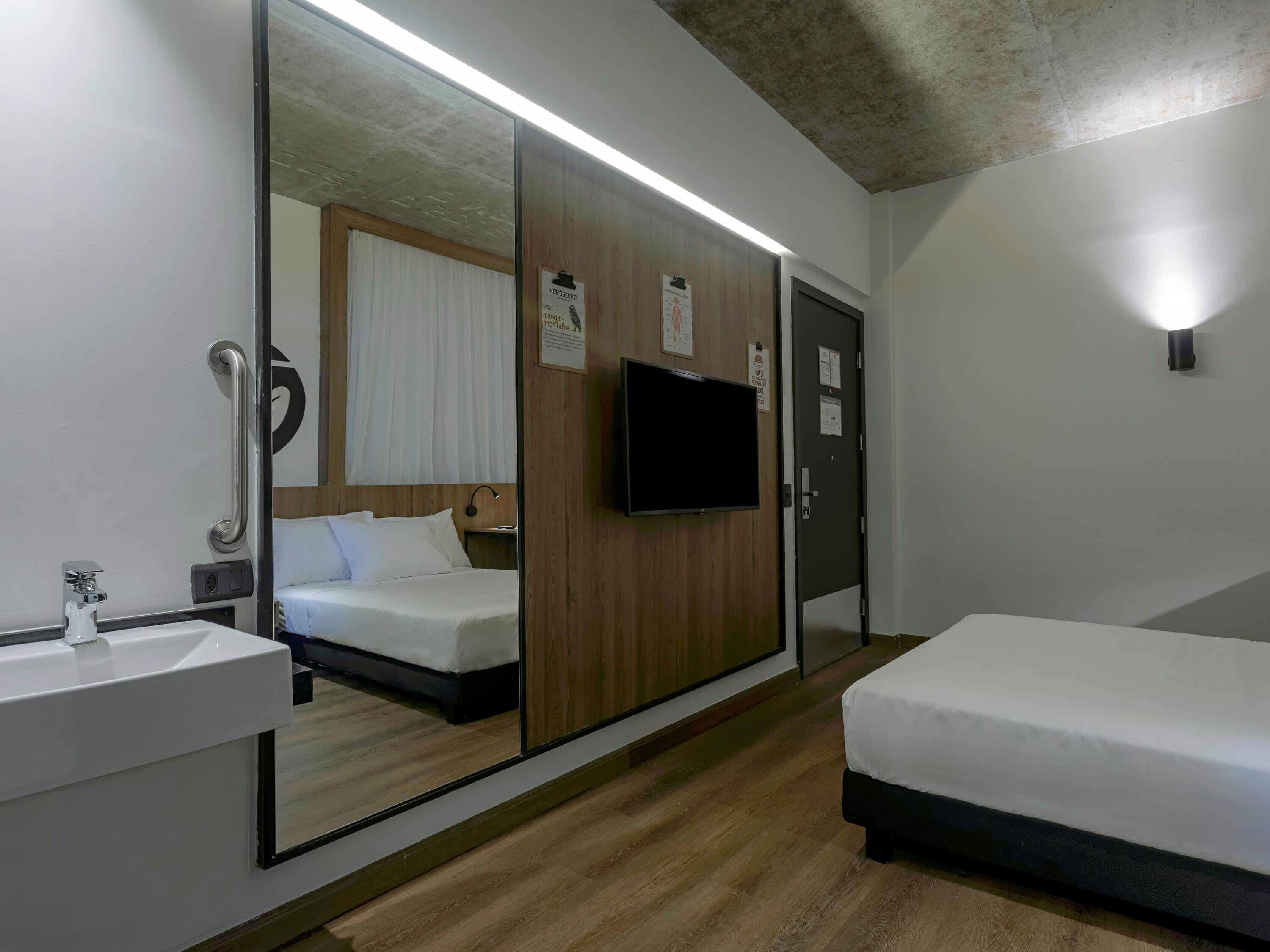 Quarto de hotel com guarda-roupa espelhado, televisão, mesa de cabeceira, pia e cama.