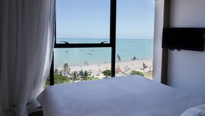 Habitación de hotel con un amplio ventanal que ofrece vistas a una playa, veleros y palmeras.