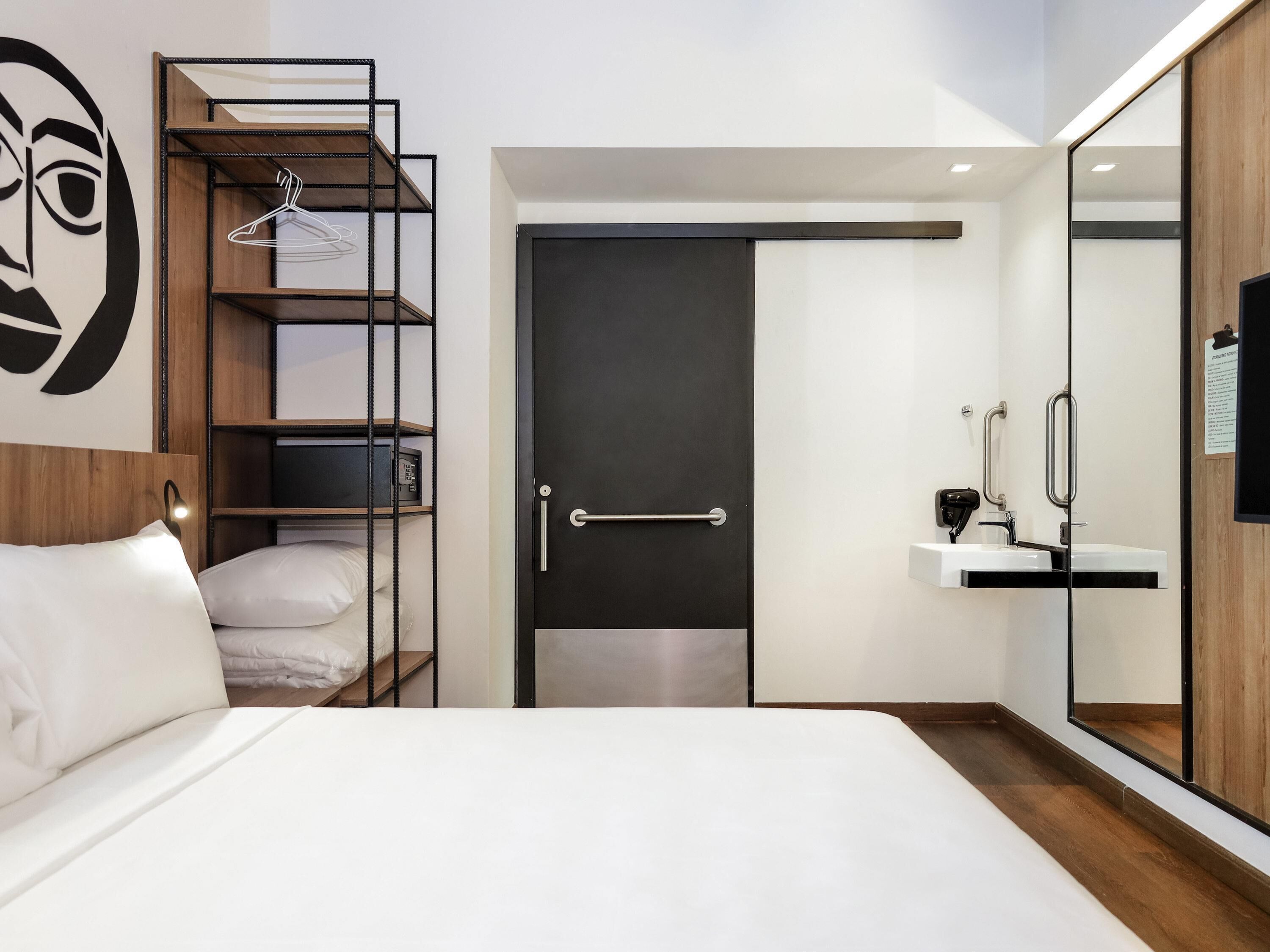 Ein modernes Hotelzimmer mit Bett, einem Schrank mit Spiegel, einem kleinen Schreibtisch und einem Regal mit aufgehängter Kleidung.
