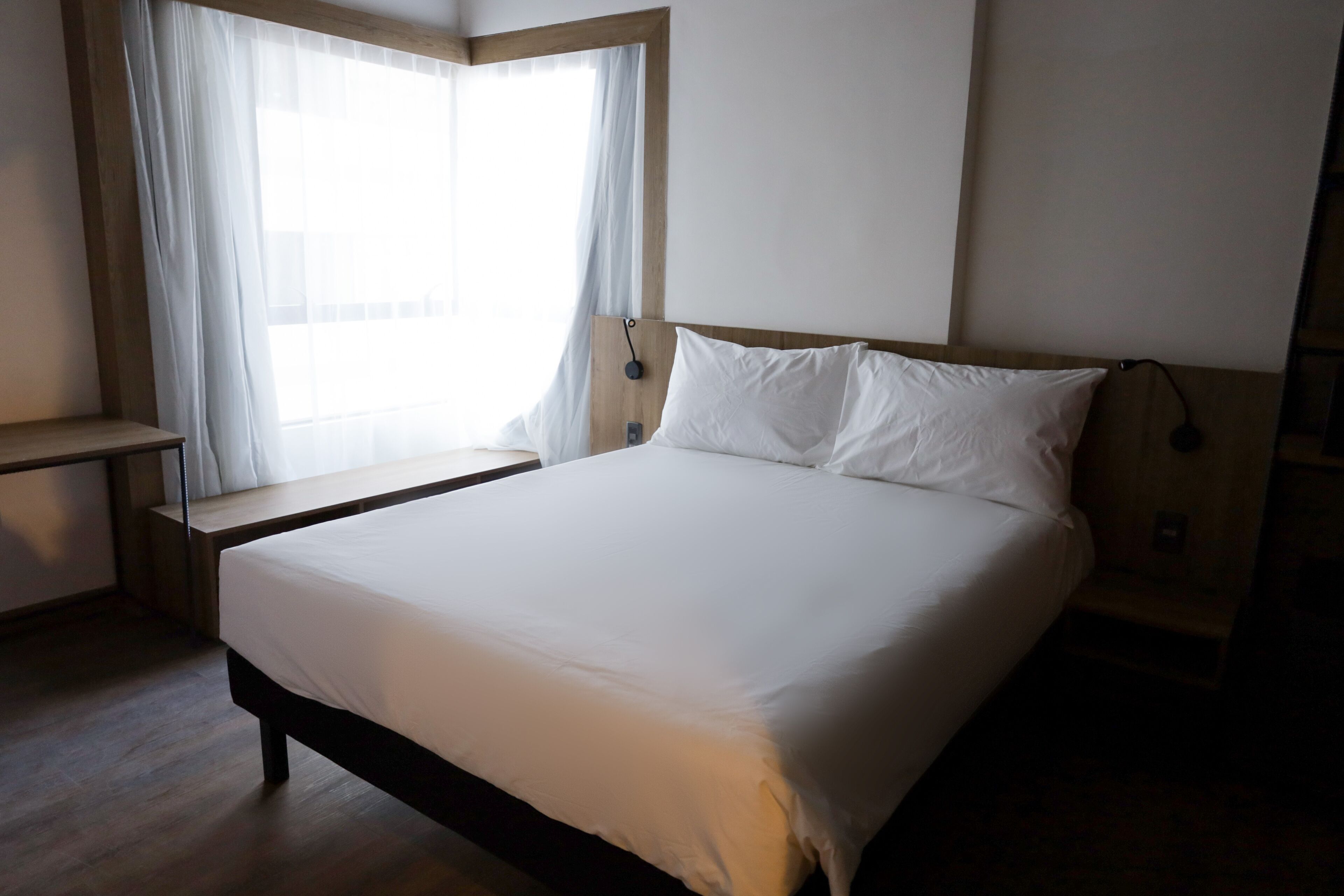 A bedroom with a large bed, a bedside table, a window with sheer curtains, and a wooden headboard.